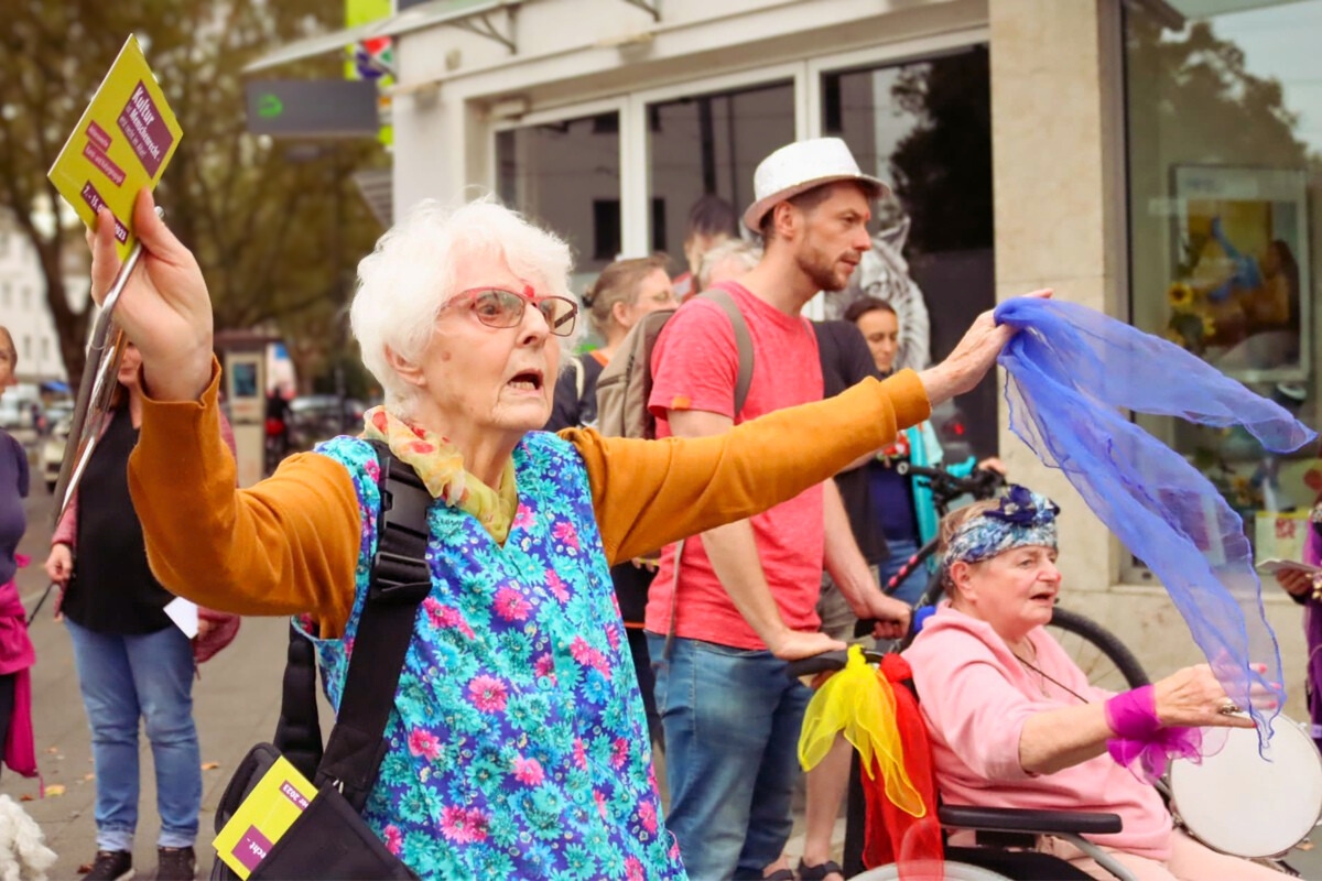Eine &auml;ltere Frau in einer gebl&uuml;mten Kittelsch&uuml;rze steht mit ausgestreckten Armen auf der Stra&szlig;e und h&auml;lt Flyer und ein violettes Tuch in den H&auml;nden. Daneben sitzt eine weitere &auml;ltere Person im Rollstuhl, der mit bunten T&uuml;chern geschm&uuml;ckt ist. Sie tr&auml;gt ein buntes Kopftuch h&auml;lt ein Tamburin in der Hand.